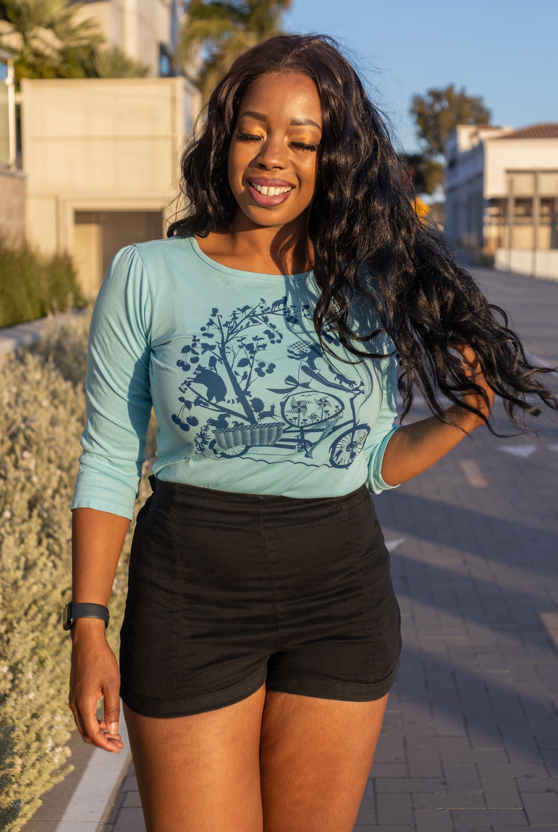 Smiling model in blue 3/4 sleeve tee with print of girl riding bicycle, worn with black shorts