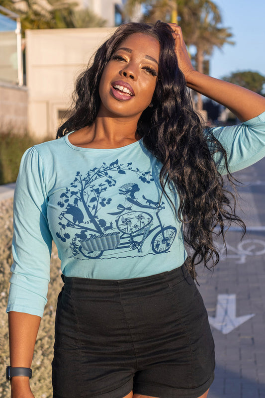 Long-haired model in blue 3/4 sleeve tee with print of girl riding bicycle with fruit trees and animals