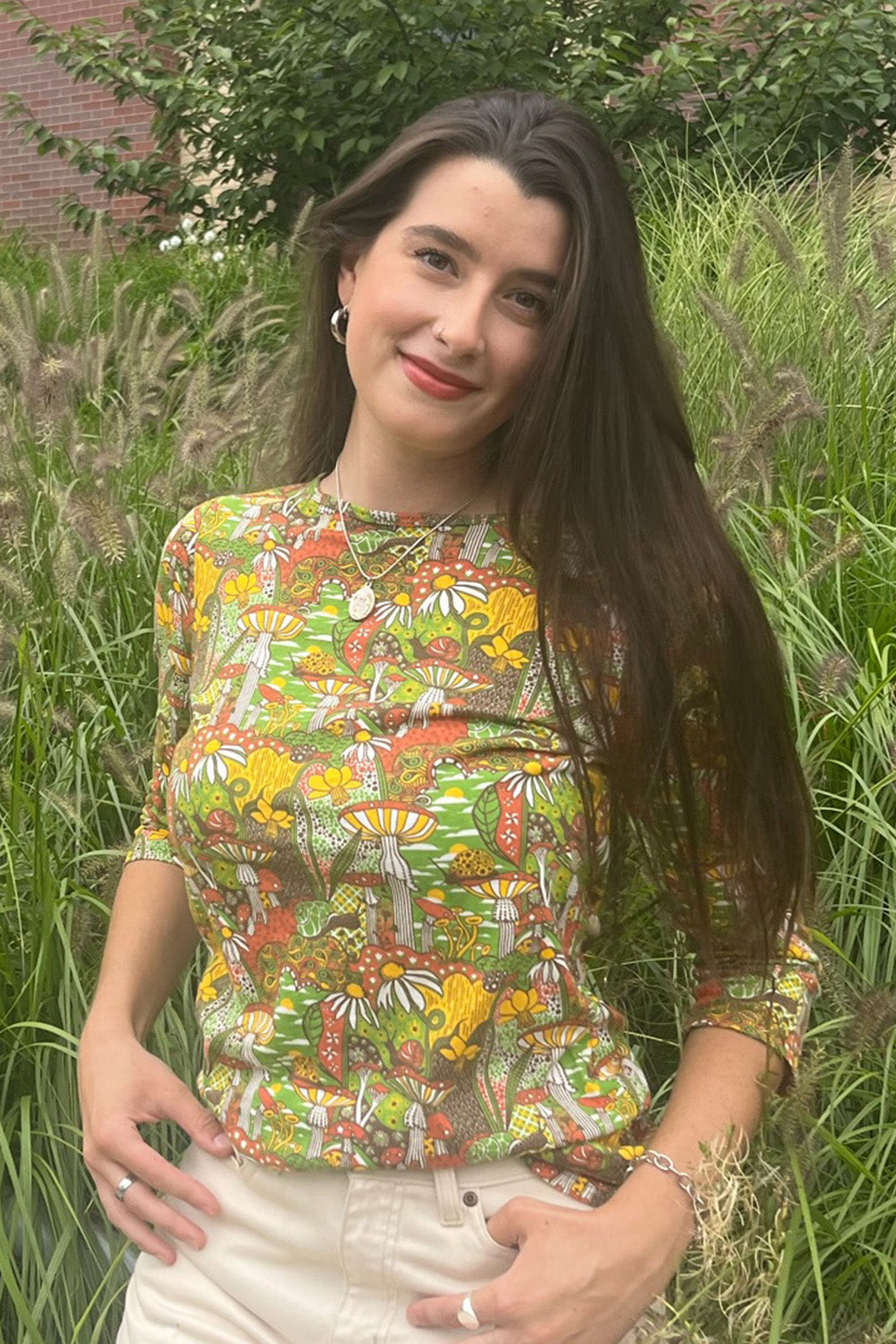 Model wearing 3/4 sleeve tee in green with fantastical landscape featuring mushrooms