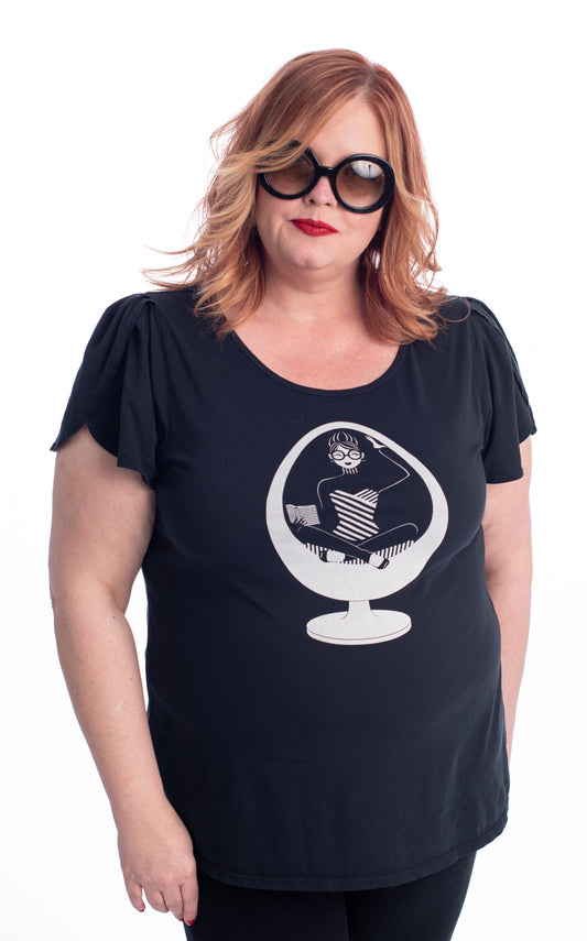Red haired model in black tee with print of girl reading in egg chair