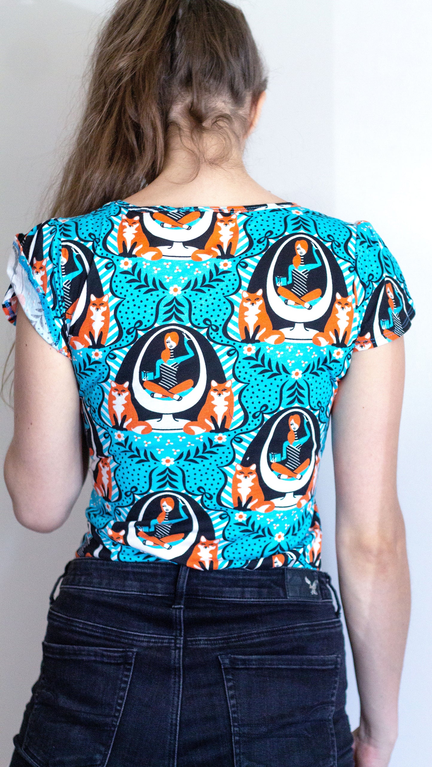 Back view of model in bright blue tulip-sleeve tee with foxes, girl reading in egg chair print