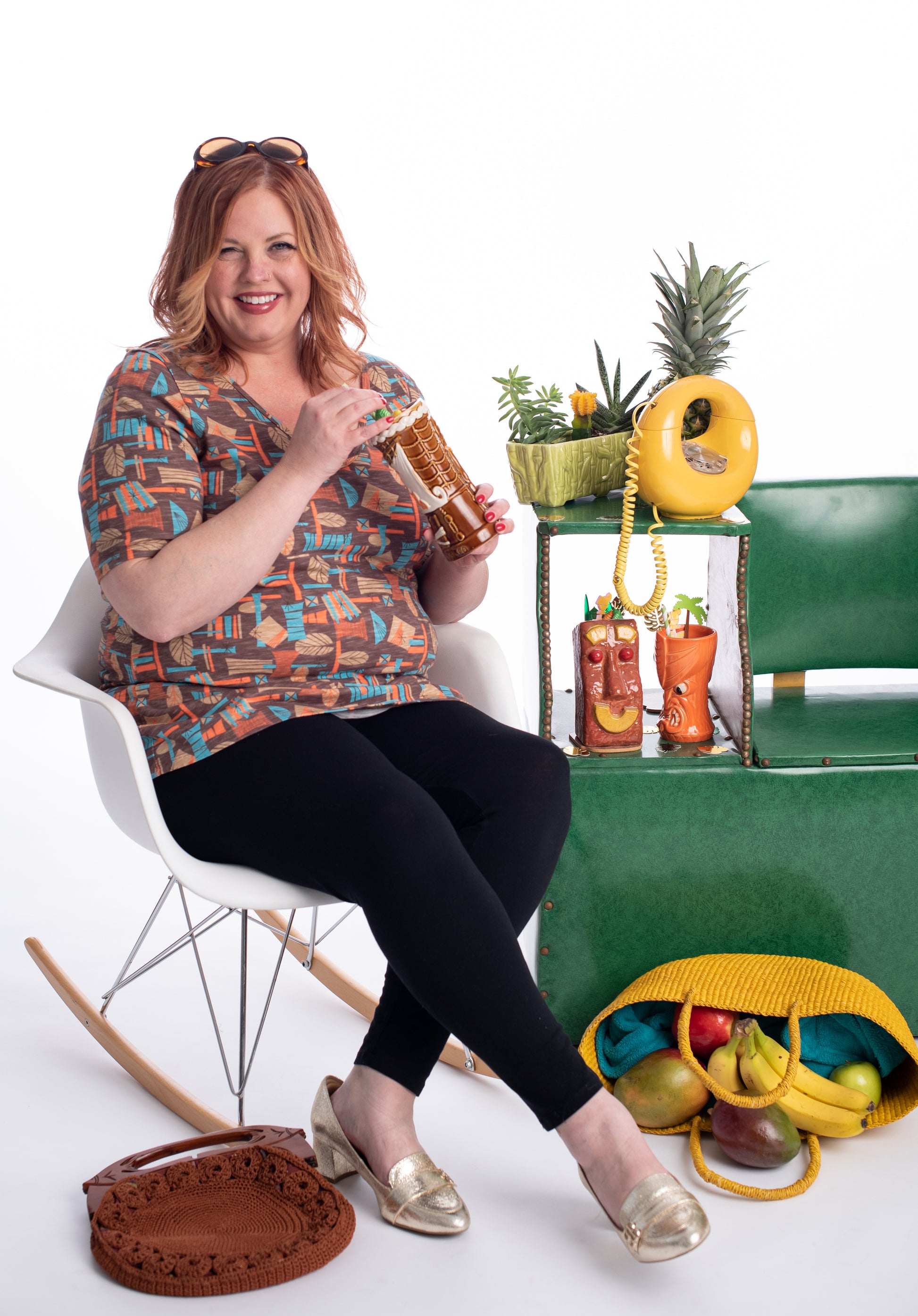 Red-headed model wearing tiki print v-neck shirt and sitting in a chair with a cocktail