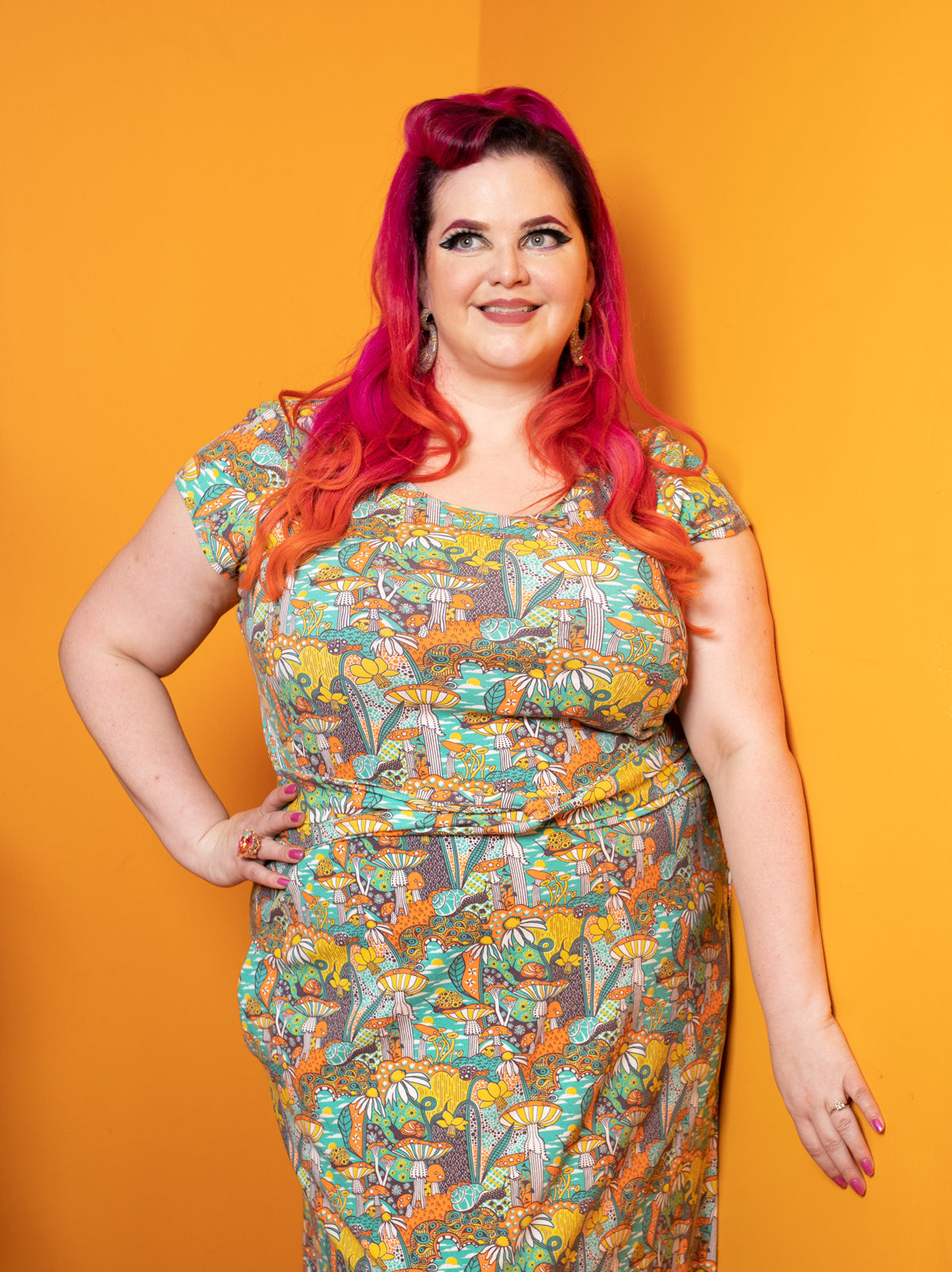 Model wearing twist belt, cap-sleeved dress featuring yellow, orange and green mushrooms, snails and flowers