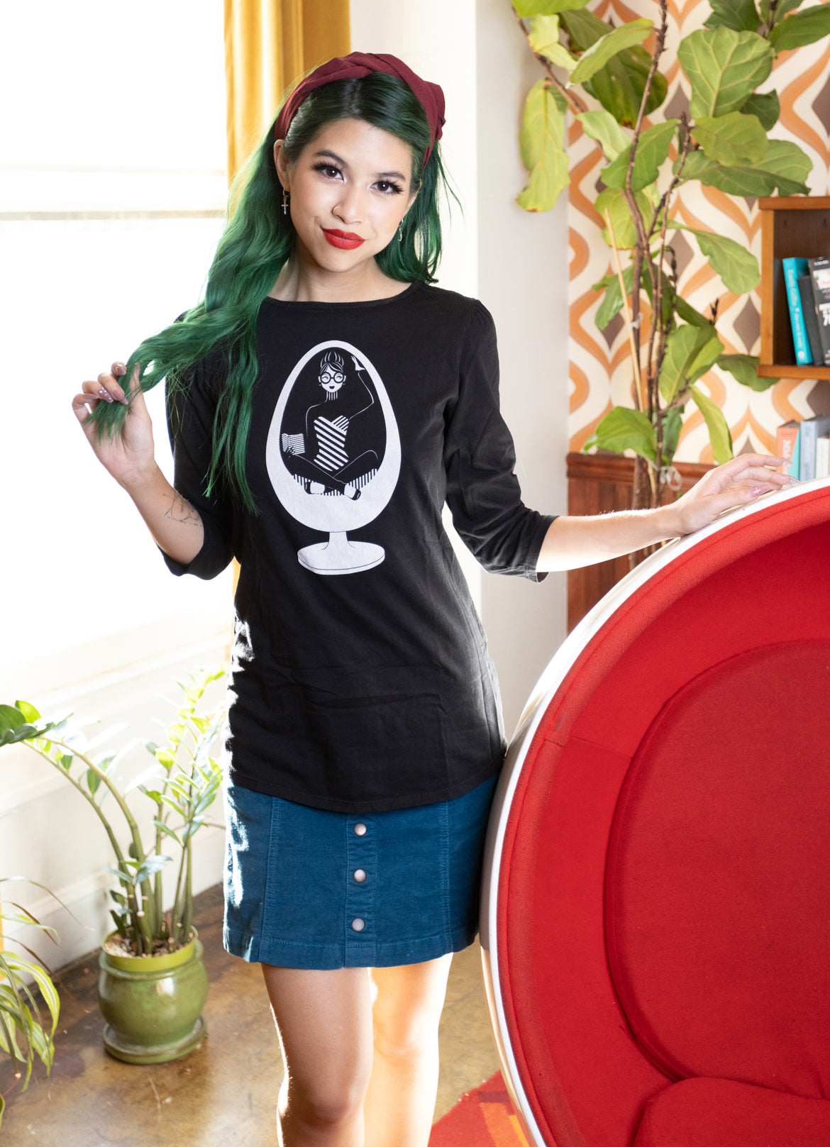 Girl with green hair standing next to ball chair and wearing black 3/4 sleeve tee with a white graphic of a girl reading in an egg chair