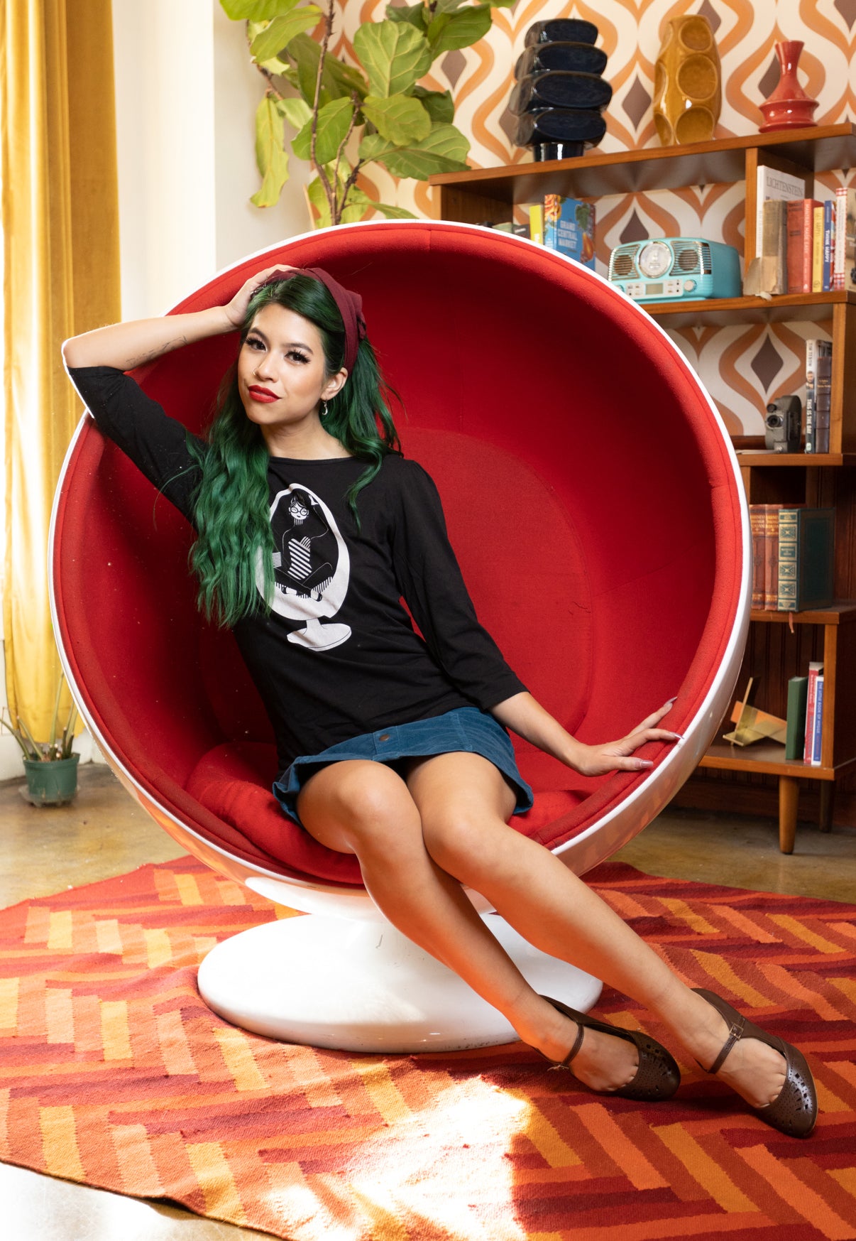 Girl with long green hair sitting in a ball chair and wearing black 3/4 sleeve tee with a white graphic of a girl reading in an egg chair