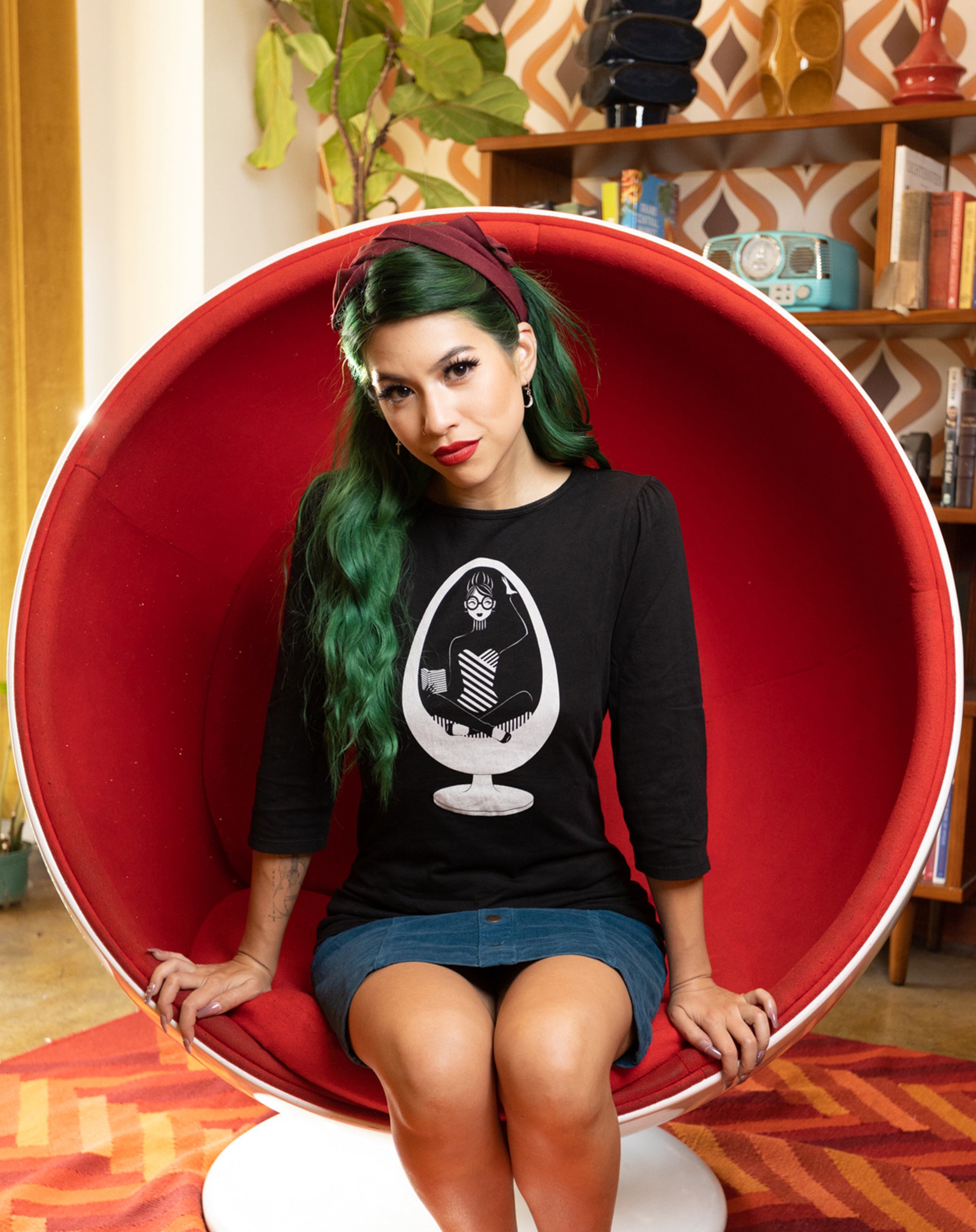 Long-haired girl sitting in ball chair wearing black 3/4 sleeve tee with white graphic of girl reading in a vintage egg chair