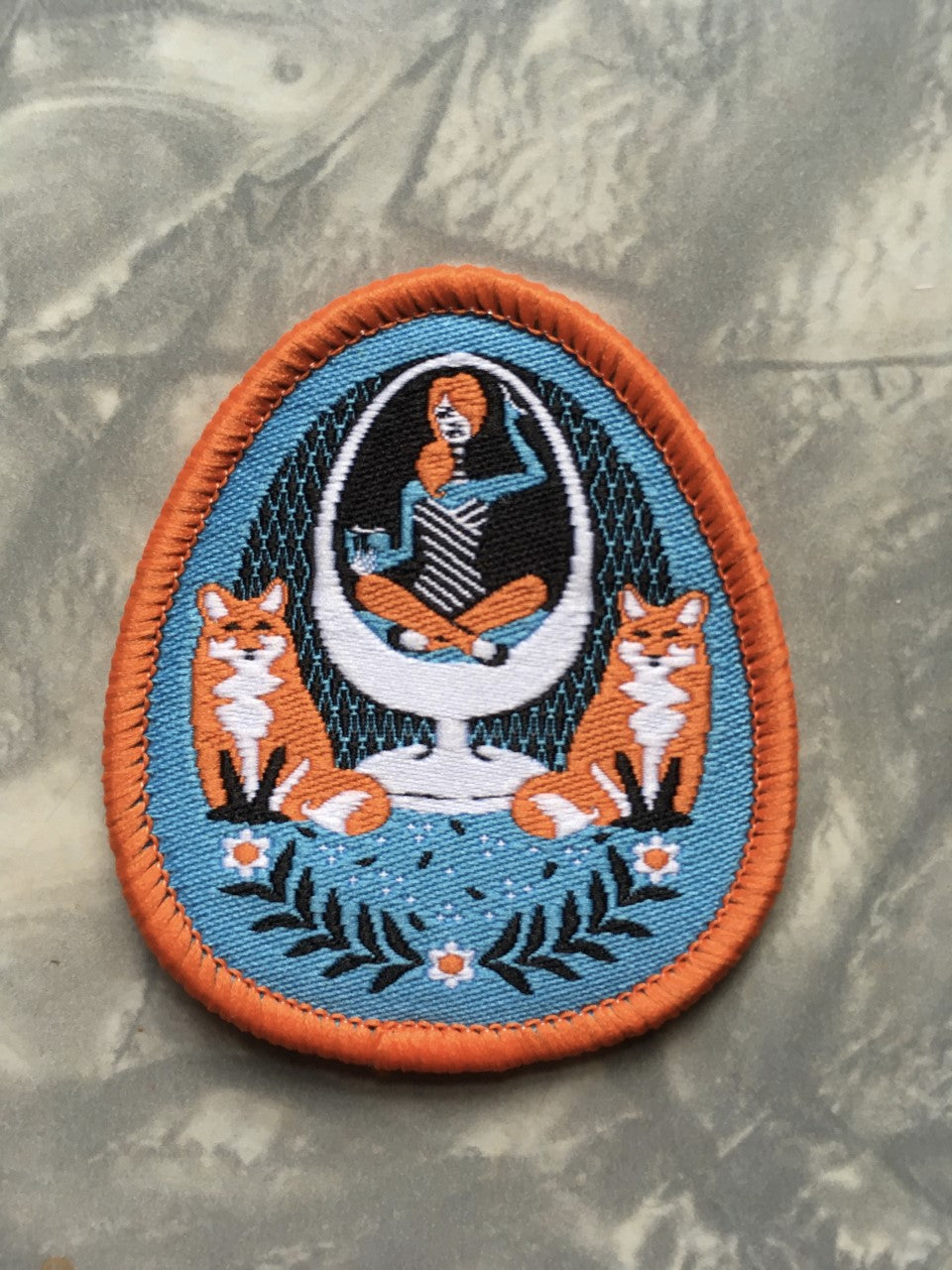 Sky blue and orange iron-on patch featuring a girl in an egg chair with 2 foxes and flower accents