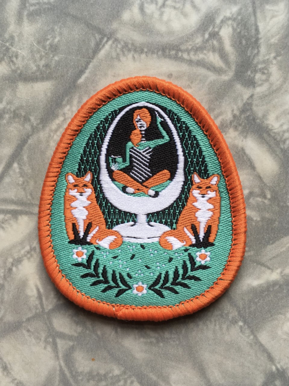 Green and orange iron-on patch featuring a girl in an egg chair with 2 foxes and flower accents