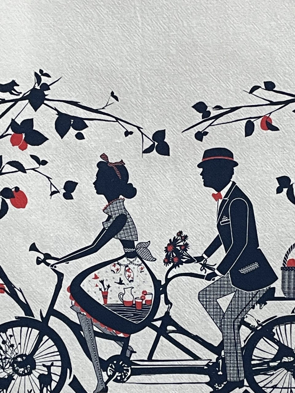Closeup of off-white tea towel with navy and red print of a couple riding a tandem among fruit trees