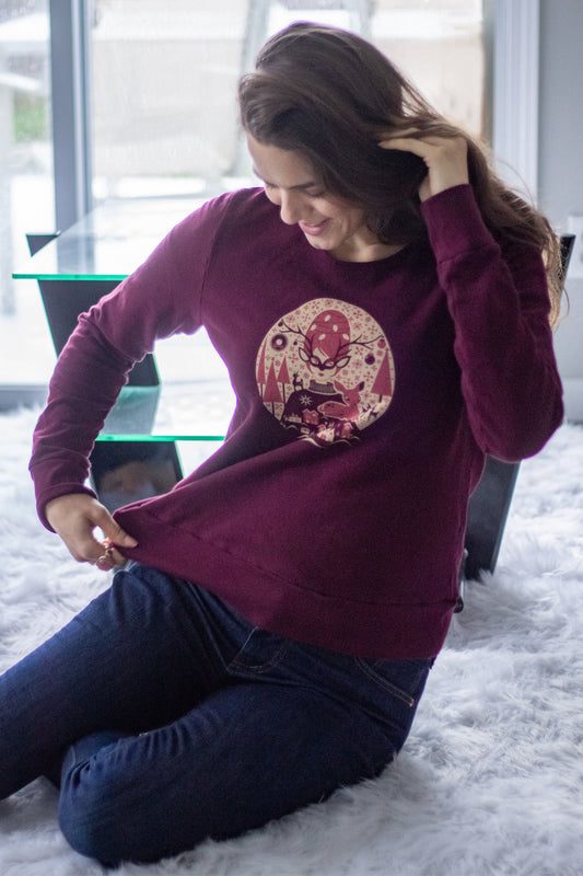 Model wearing plum sweatshirt with red and white graphic of Christmas girl, reindeer, and pine trees