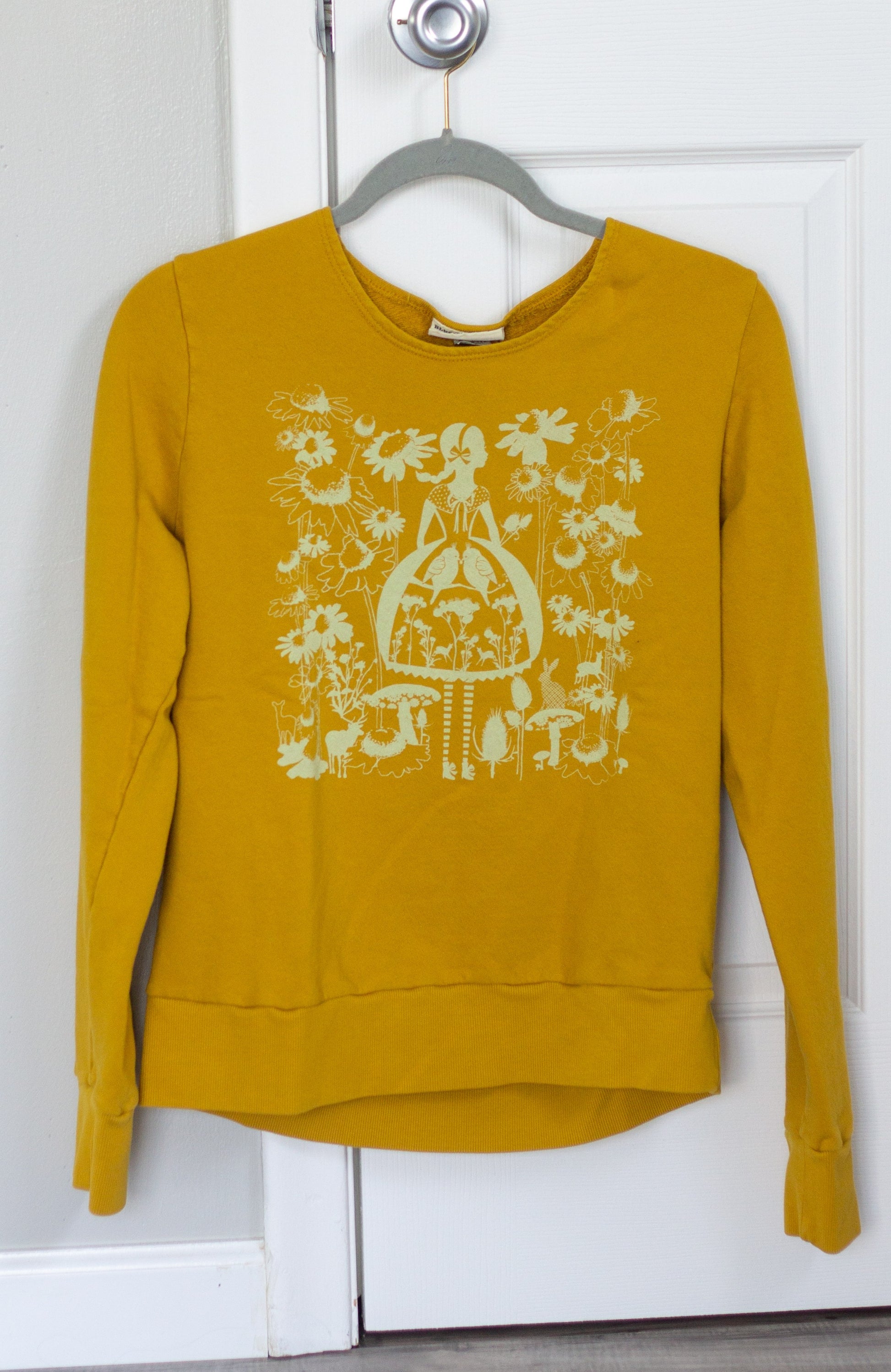 Yellow sweatshirt on hanger with white screen print of cute girl surrounded by flora and fauna 