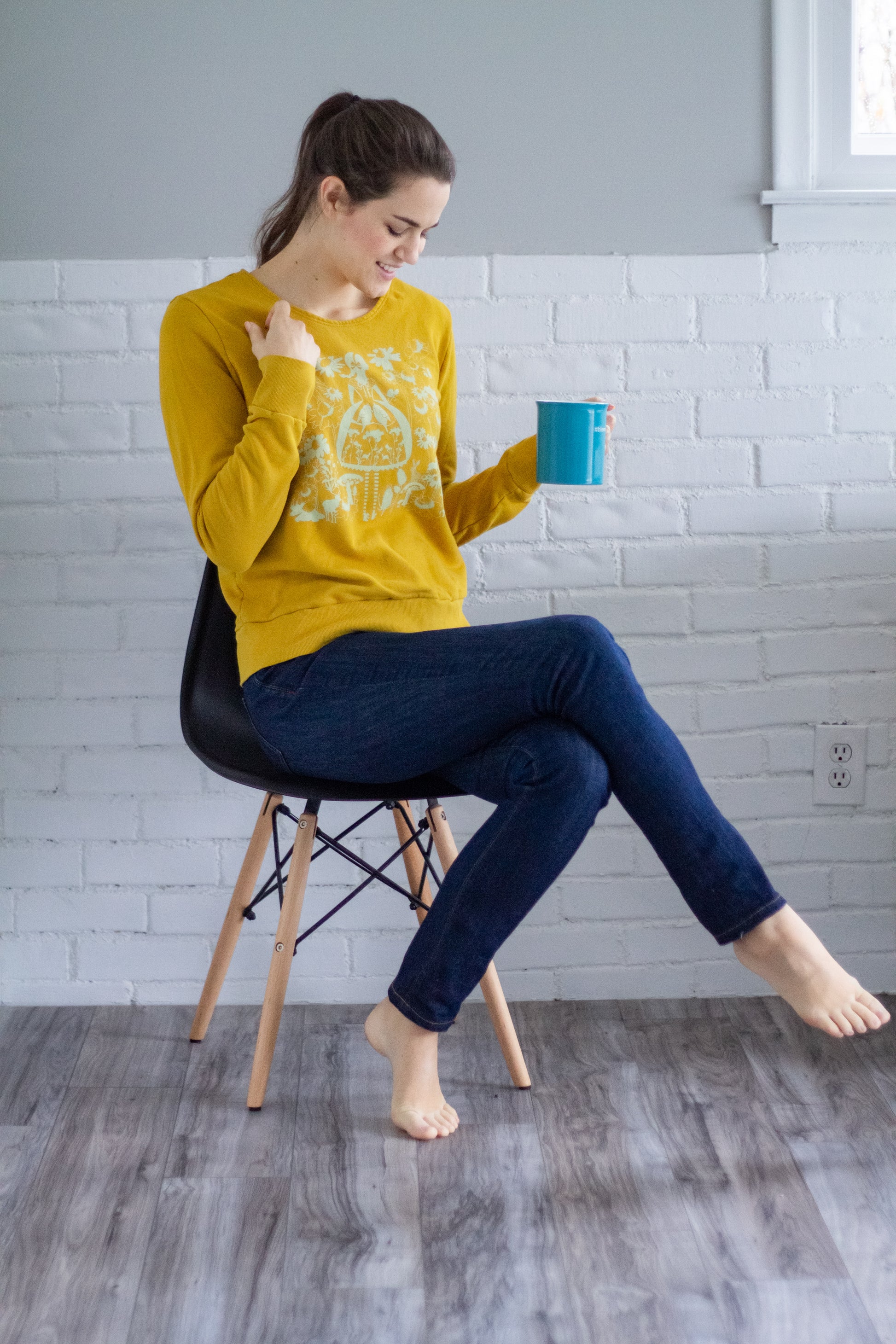 Model seated with coffee mug wearing sweatshirt with white screen print of cute girl surrounded by flora and fauna
